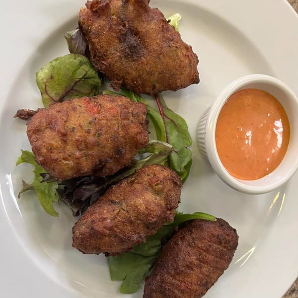 Four crispy brown Caribbean fritters on a bed of greens with a side of orange dipping sauce.
