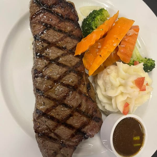 Grilled steak with grill marks, mashed potatoes, carrots, broccoli, and a side of brown sauce.